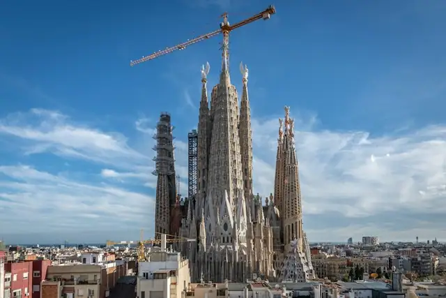 A large basilica under construction with several tall spires and a crane on top, set against a clear blue sky.