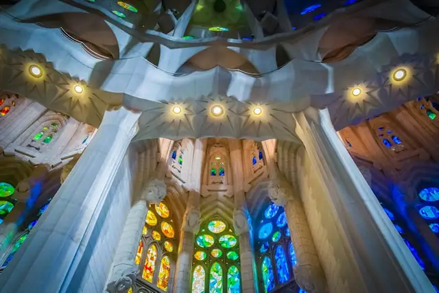 Interior of a cathedral with illuminated stained glass windows in various colors and ornate architectural details under ceiling lights.