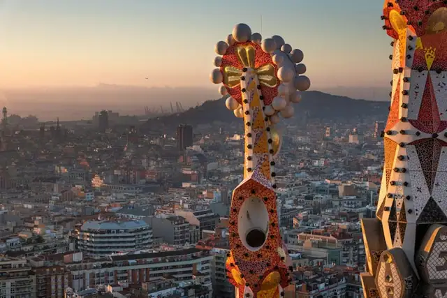 A colorful, ornate spire adorned with mosaic tiles towers over a densely built cityscape at sunset.