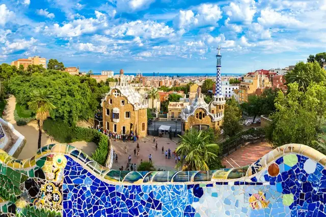 Colorful mosaic foreground with whimsical buildings, lush trees, and numerous people. A cityscape and cloudy sky in the background.